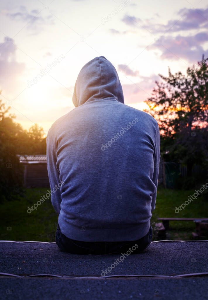 Rear View of a Lonely Young Man in a Hoodie sit on the Sunset Background Outdoor