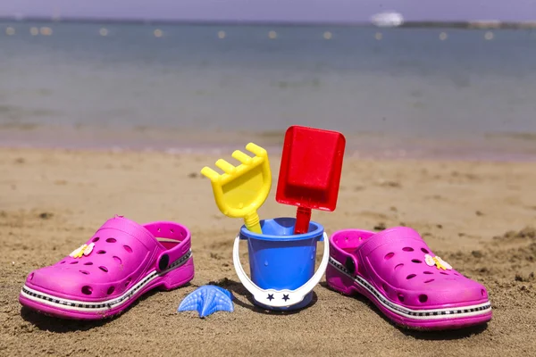 Crocs de praia rosa e brinquedos de areia azul na praia arenosa. Chinelos de praia em primeiro plano e mar borrado no fundo — Fotografia de Stock