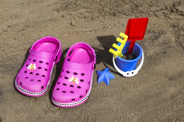 Pink beach crocs and blue sand toys on sandy beach.Beach flip flops in the foreground and blurred sea in the background — Stock Photo, Image