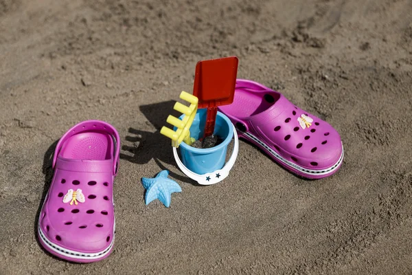 Crocs de praia rosa e brinquedos de areia azul na praia arenosa. Chinelos de praia em primeiro plano e mar borrado no fundo — Fotografia de Stock