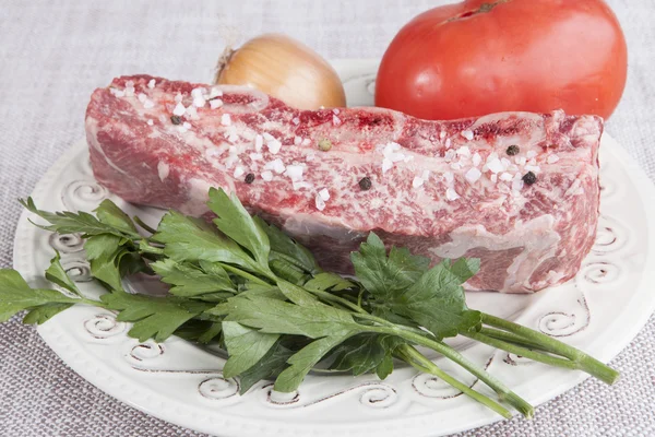 Um pedaço de carne fresca com sal marinho e pimenta preta, tomate, cebola em um prato de porcelana — Fotografia de Stock