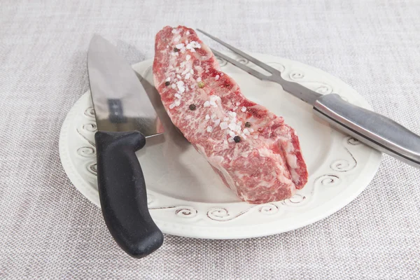 A piece of fresh marbled beef with sea salt and black pepper, knife and fork on a porcelain plate — 스톡 사진