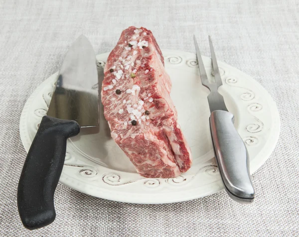 Un trozo de ternera fresca de mármol con sal marina y pimienta negra, cuchillo y tenedor en un plato de porcelana —  Fotos de Stock