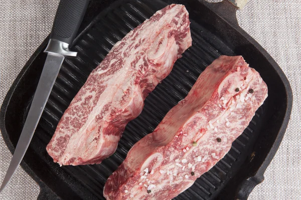 Primer plano de dos trozos de ternera fresca de mármol con sal marina y pimienta negra, cuchillo en una parrilla de hierro fundido —  Fotos de Stock