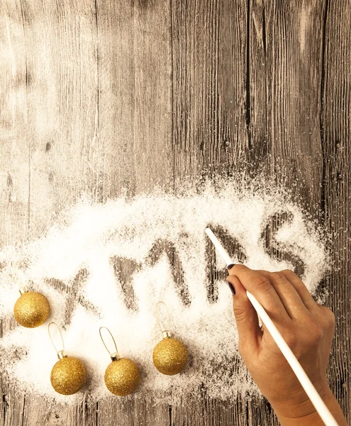 Christmas card with a female hand writing xmas, gold balls in the snow on a wooden background. — Stock Fotó