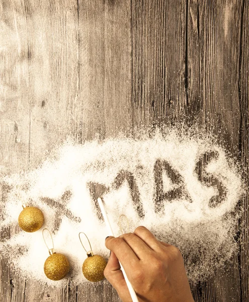 Christmas card with a female hand writing xmas, gold balls in the snow on a wooden background. — Stock Fotó