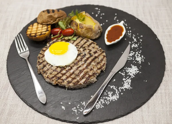 Big juicy grilled steak marbled beef with egg baked potatoes with barbecue sauce. Served on a stone plate with a fork and spoon on a textile background.