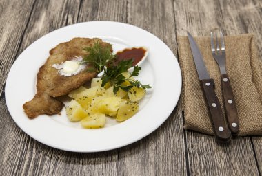 Petite Wiener schnitzel with boiled potatoes and ketchup. Served on a white porcelain plate with fork and knife on a wooden background.