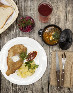 Business lunch : Petite Wiener schnitzel with boiled potatoes and ketchup. Served on a white porcelain plate with fork and knife on a wooden background. Beet  salad and tomato, red pepper soup, sauce 