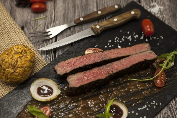 Bife Ribeye de carne de vaca de mármore com legumes e molho de churrasco. Servido em uma placa de pedra preta com garfo e faca  . — Fotografia de Stock