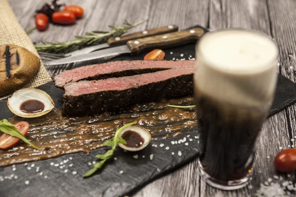 Ribeye steak z mramorové hovězí maso se zeleninou a barbecue omáčkou. Podávané na talíř z černého kamene s vidličkou a nožem a pivo — Stock fotografie