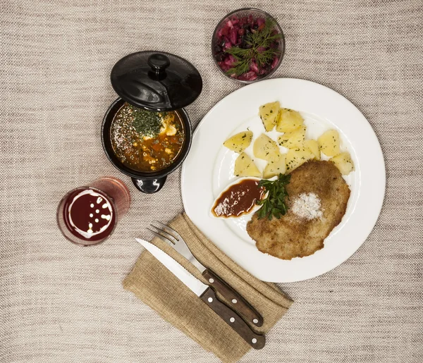 Business lunch : Petite Wiener schnitzel with boiled potatoes and ketchup. Served on a white porcelain plate with fork and knife on a wooden background. Beet salad and tomato, red pepper soup, sauce — Stock Image Business lunch : Petite Wiener schnitzel with boiled potatoes and ketchup. Served on a white porcelain plate with fork and knife on a wooden background. Beet salad and tomato, red pepper soup, sauce Stock Picture