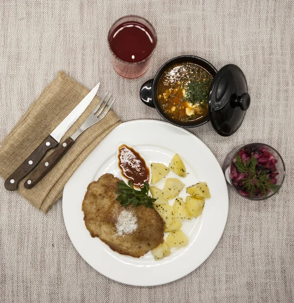 Business lunch : Petite Wiener schnitzel with boiled potatoes and ketchup. Served on a white porcelain plate with fork and knife on a wooden background. Beet salad and tomato, red pepper soup, sauce — Stock Image Business lunch : Petite Wiener schnitzel with boiled potatoes and ketchup. Served on a white porcelain plate with fork and knife on a wooden background. Beet salad and tomato, red pepper soup, sauce Stock Image