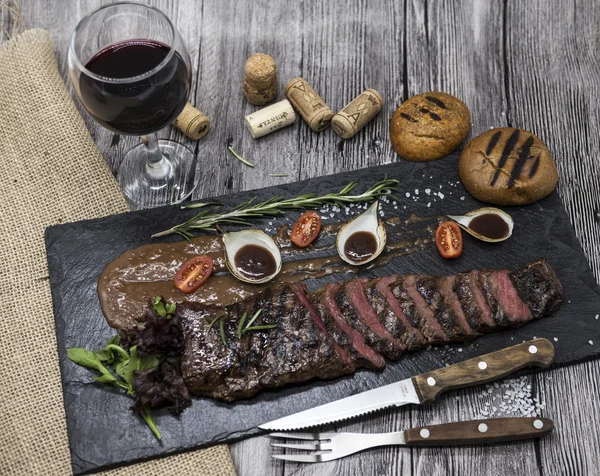 Delicioso y jugoso rosbif caliente cortado en deliciosos trozos de carne y verduras. Servido en un plato de piedra negra con un tenedor y cuchillo y pimienta . —  Fotos de Stock