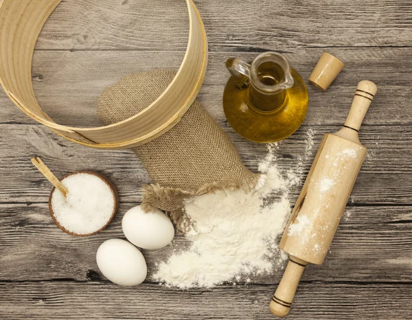 Wheat flour in a canvas bag,sieve, the olive oil in a glass carafe, a large salt shaker wood, raw eggs, a wooden rolling pin: set for making homemade bread dough on a beautiful dark wooden background — Stockfoto