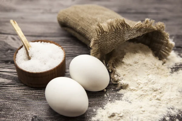 Farina di frumento in un sacchetto di tela, un grande legno di agitatore di sale, uova crude: set per fare pasta di pane fatta in casa su un bellissimo sfondo di legno scuro — Foto Stock