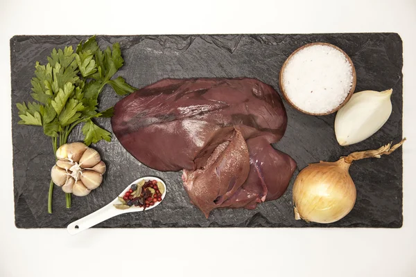 Pieces of fresh raw beef liver, onion, garlic, spices, salt on the stone plate on a white background. — Stock Photo, Image