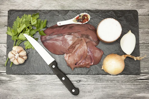 Pieces of fresh raw beef liver, onion, garlic, spices, salt, knife on the stone plate on a white background. — Stock Fotó