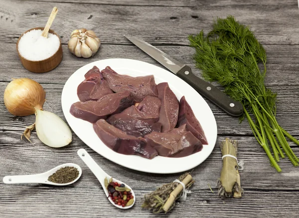 Pieces of fresh raw beef liver, onion, garlic, spices, dill, parsley, salt, knife, olive oil on porcelain plate on a wooden background. — ストック写真