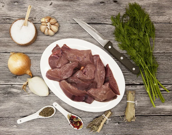 Pieces of fresh raw beef liver, onion, garlic, spices, dill, parsley, salt, knife, olive oil on porcelain plate on a wooden background. — Zdjęcie stockowe