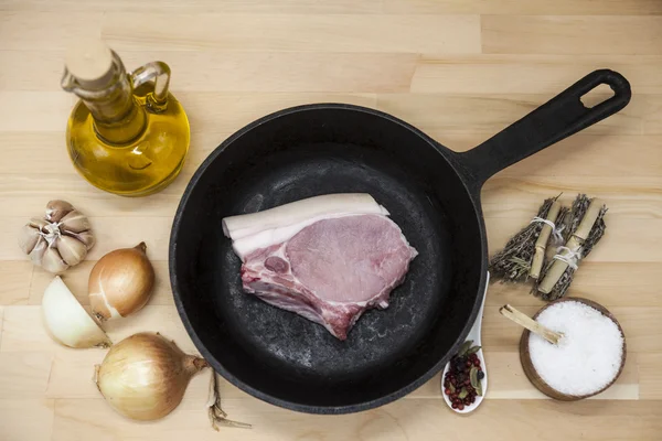 Kawałek pyszne świeże surowy wieprzowina zbliżenie na patelni żeliwnych, cebula, czosnek, przyprawy, sól, oliwy z oliwek na stole w kuchni rustykalny. — Zdjęcie stockowe