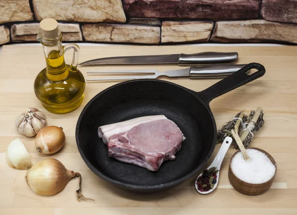 Un pedazo de delicioso cerdo crudo fresco de cerca en una sartén de hierro fundido, cebollas, ajo, especias, sal, aceite de oliva, tenedor, cuchillo en una mesa de cocina rústica — Foto de Stock