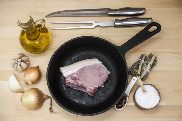 Kawałek pyszne świeże surowy wieprzowina zbliżenie na patelni żeliwnych, cebula, czosnek, przyprawy, sól, oliwy z oliwek, widelec, nóż na stole rustykalna kuchnia — Zdjęcie stockowe