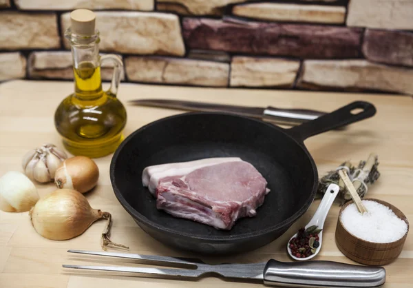 Un pedazo de delicioso cerdo crudo fresco de cerca en una sartén de hierro fundido, cebollas, ajo, especias, sal, aceite de oliva, tenedor, cuchillo en una mesa de cocina rústica — Foto de Stock