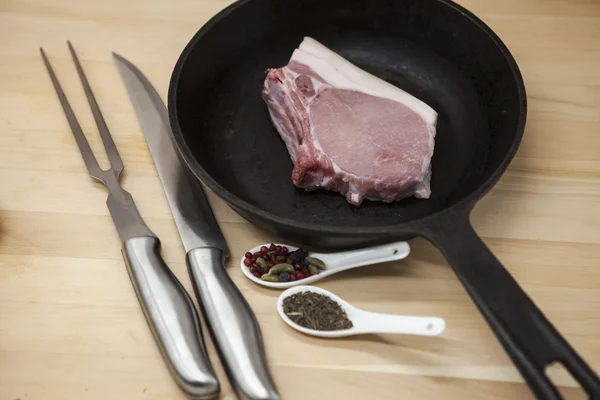 Egy darab finom, friss, nyers sertéshús közeli villát, kést, fűszer, hagyma, fokhagyma, egy öntöttvas serpenyőben a táblázat rusztikus konyha. — Stock Fotó