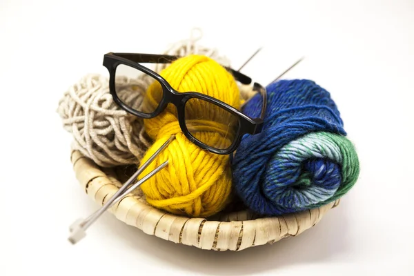 Três bolas de fios de lã, amarelo, azul, bege e aço agulhas de tricô e óculos em uma cesta de madeira em um fundo branco . — Fotografia de Stock