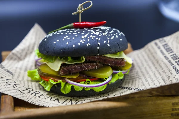 Black burger. A burger with a black roll slices of juicy marble beef, fused cheese, fresh salad and sauce of a barbecue. A burger on the newspaper on a wooden tray  on a dark background. — Stockfoto