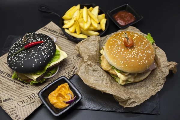 Set des schwarzen Burgers und des klassischen amerikanischen Burgers. Black Burger Roll Scheiben saftigen Marmor Rindfleisch, geschmolzenem Käse, frischem Salat mit Pommes frites auf einem gusseisernen Grill, mit Chili-Sauce und Guakomol — Stockfoto