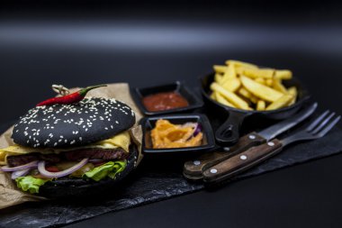Set of the black burger. Stone plate with: black burger roll slices of juicy marble beef, fused cheese, fresh salad with French fries on a cast-iron griddle, with chili sauce and guakomole.