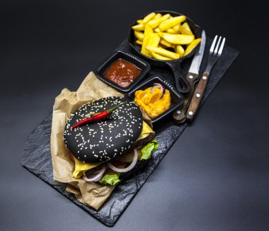 Set of the black burger. Stone plate with: black burger roll slices of juicy marble beef, fused cheese, fresh salad with French fries on a cast-iron griddle, with chili sauce and guakomole.