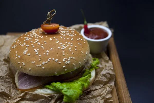 Klassische amerikanische frische saftige Burger mit Huhn und Schinken auf einem hölzernen Tablett mit einer würzigen Chilisoße. schönes Foto auf dunklem Hintergrund — Stockfoto