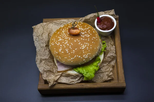 Hambúrguer suculento americano clássico com frango e presunto em uma bandeja de madeira com um molho picante de pimenta. Bela foto em um fundo escuro — Fotografia de Stock