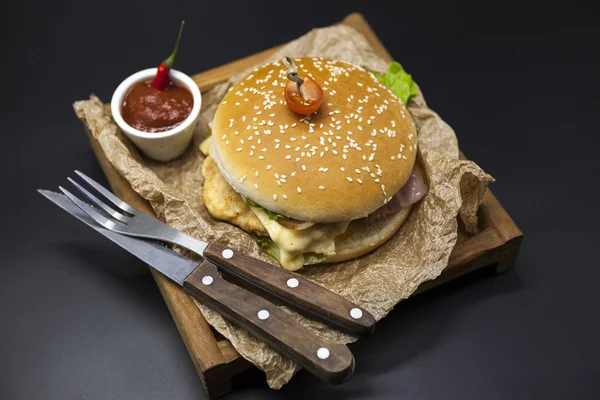 Classical American fresh juicy burger with chicken and ham on a wooden tray with a spicy chili sauce. Beautiful photo on a dark background — Stockfoto