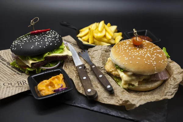 Set of the black burger and classical American burger . Black burger roll slices of juicy marble beef, fused cheese, fresh salad with French fries on a cast-iron griddle, with chili sauce and guakomol — Zdjęcie stockowe