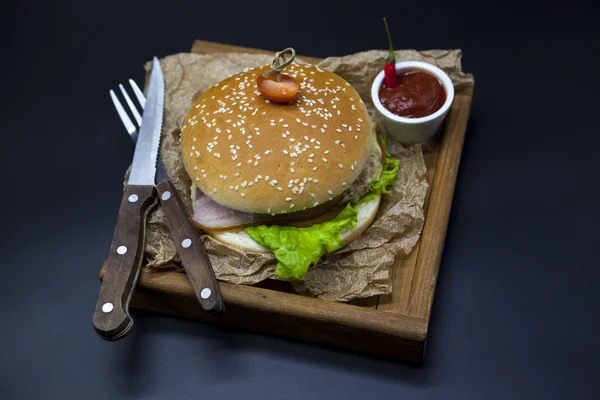 Hambúrguer suculento americano clássico com frango e presunto em uma bandeja de madeira com um molho picante de pimenta. Bela foto em um fundo escuro — Fotografia de Stock