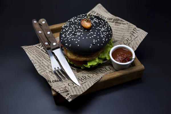 Schwarzburger. Ein Burger mit einer schwarzen Rolle Scheiben saftigen Marmor Rindfleisch, geschmolzenem Käse, frischem Salat und Sauce von einem Grill. ein Burger auf einem hölzernen Tablett mit Messer und Gabel auf einem dunklen Ba — Stockfoto