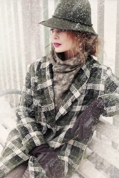 在雪地里 一个戴着复古帽子 穿着格子呢外套 站在长椅上的漂亮女人的画像 免版税图库照片