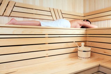 Beyaz havluya sarılı sıska bir kadın geleneksel saunada bankta yatıyor.