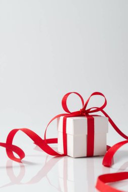 Üzerinde kırmızı saten bir kurdele bulunan küçük bir kare beyaz hediye kutusu Noel, yeni yıl ya da doğum günü teması için mükemmel bir fiyonk oluşturuyor. Kurdele, kutunun etrafında yansıtıcı beyaz bir yüzey etrafında dönüyor.