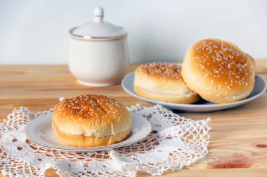 Beyaz tabakta susamlı taze hamburger çörekleri ve tahta masada beyaz şeker kasesi. Seçici odak.