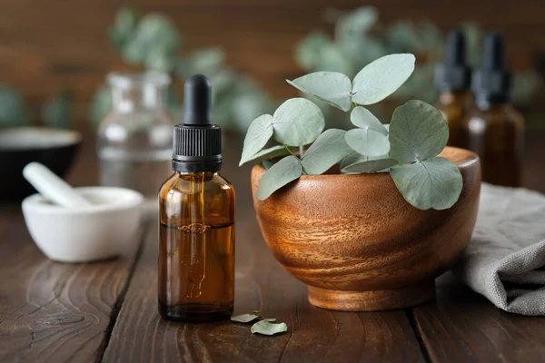 Dropper bottle of eucalyptus essential oil and wooden bowl of green eucalyptus leaves. Mortar and oil bottles on background, not in focus.