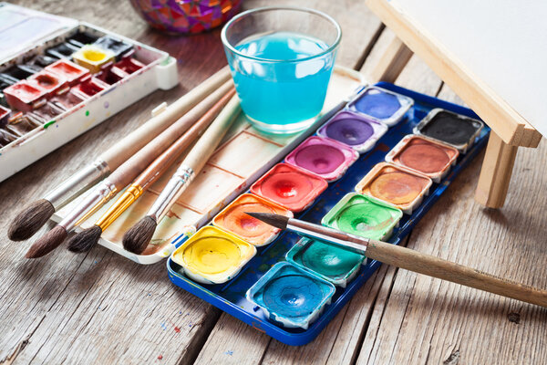 Box of watercolor paints, art brushes, glass of water and easel 