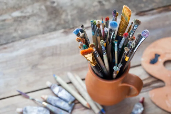 Paintbrushes in a jug from potters clay, palette and paint tubes — Stock Photo, Image