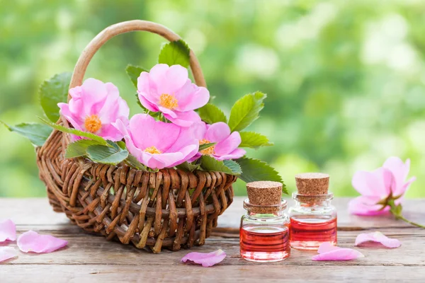 Rustic wicker basket with pink rose hip flowers and bottles of e — Stock Photo, Image