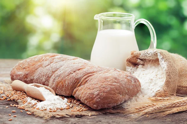 Pane fresco, brocca di latte, sacco di farina e spighe di grano su legno — Foto Stock