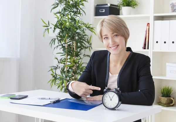Business woman, at the table, shows the clock and smiling — Stock Photo, Image